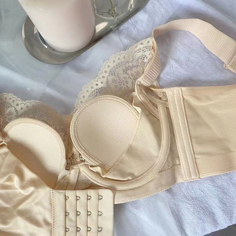 A close-up view of a beige lace bra with soft padded cups and adjustable wide straps. The inner side of the bra is shown, featuring a removable padding insert and detailed lace along the top edge of the cups. The back includes a multi-row hook-and-eye closure for adjustable support. The bra is laid on a light fabric next to a candle on a metal tray.